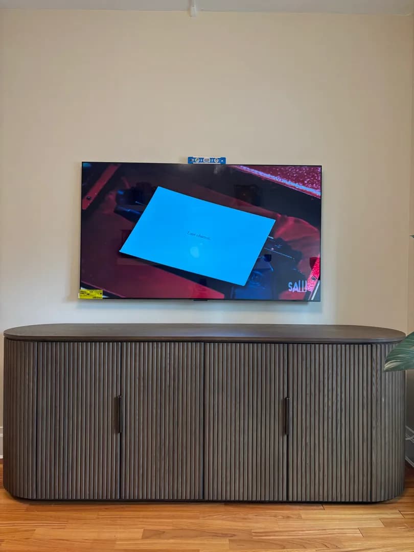A TV mounted flat on a cream wall above a long dark ribbed console, with a small level resting on top.