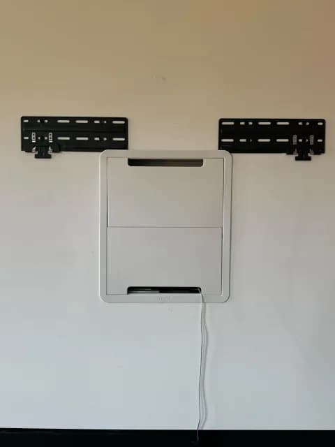 A closed white in-wall AV box between two black TV mount brackets on a wall, with a cable hanging below.