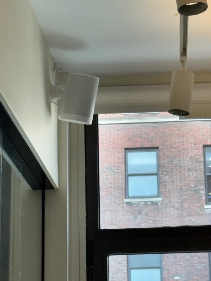 A white Sonos speaker mounted on a bracket high on the wall beside a window.