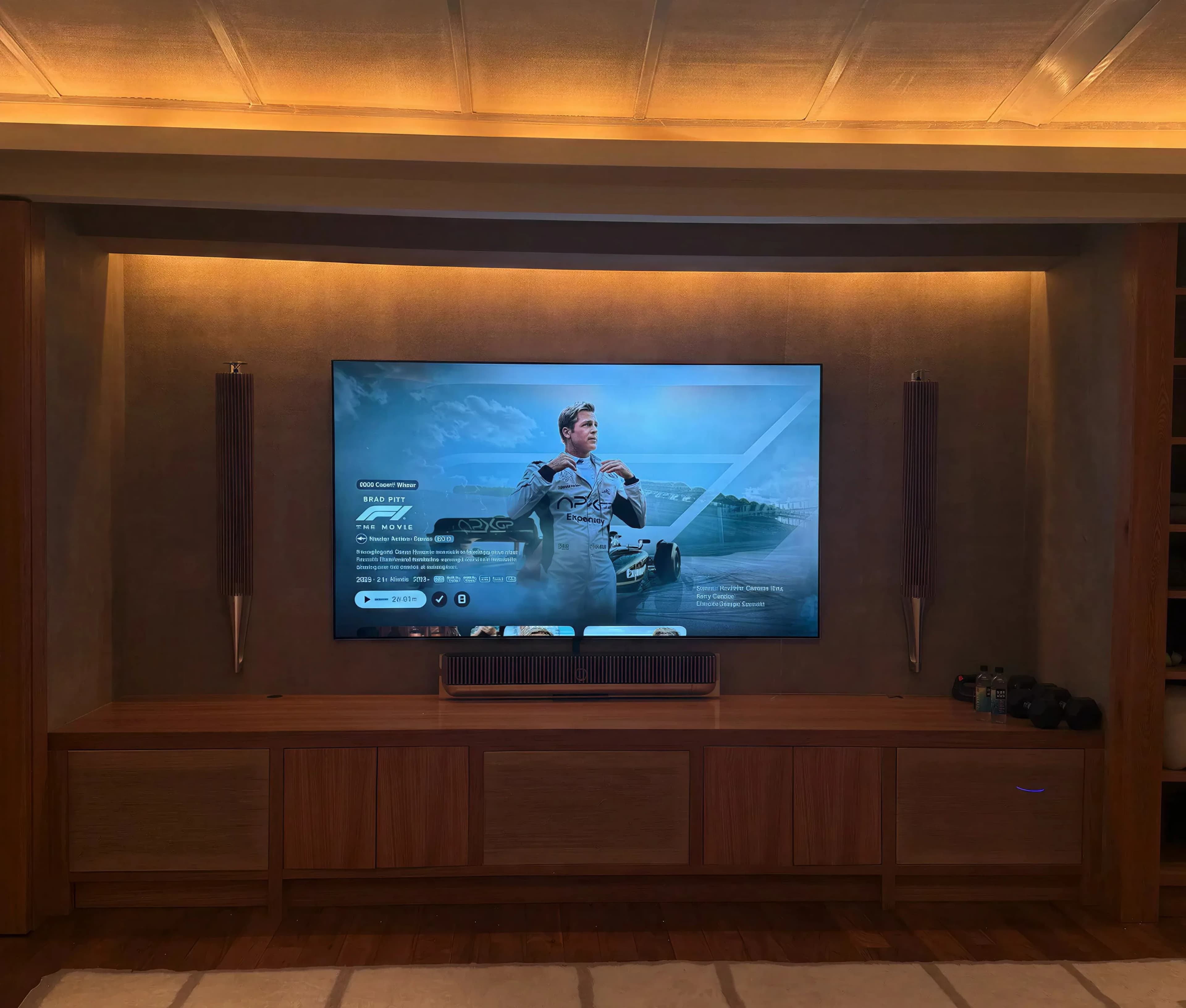 A large TV on a walnut built-in TV wall with Bang & Olufsen speakers and a soundbar on a built-in console, in a cove-lit home theatre
