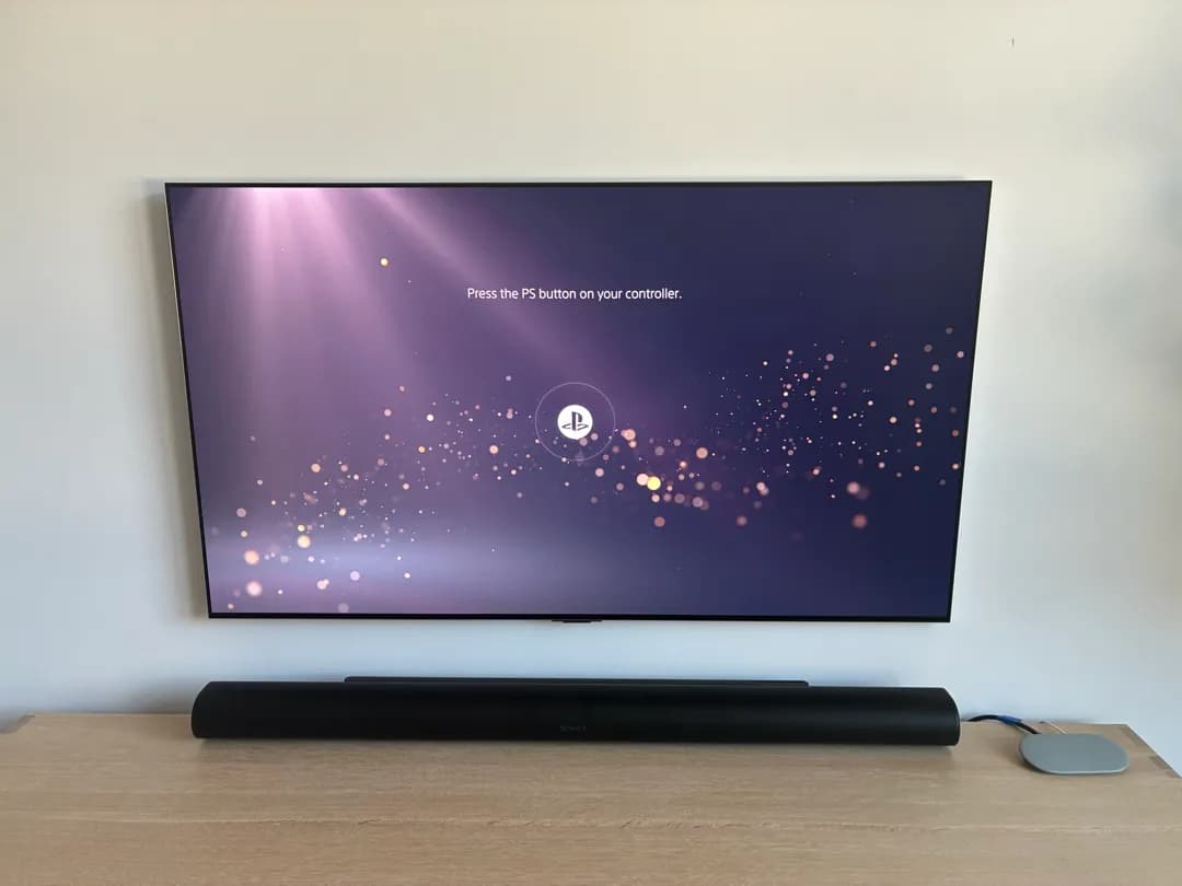 A wall-mounted TV displays a PlayStation controller connect screen above a soundbar on a wood console.