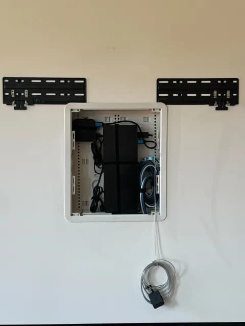 An open in-wall AV cable box with equipment inside, flanked by two black TV mount brackets on a wall.