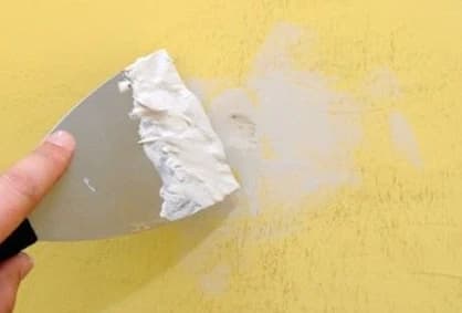 Hand using a putty knife to patch and smooth wall anchor holes on a yellow wall after dismounting
