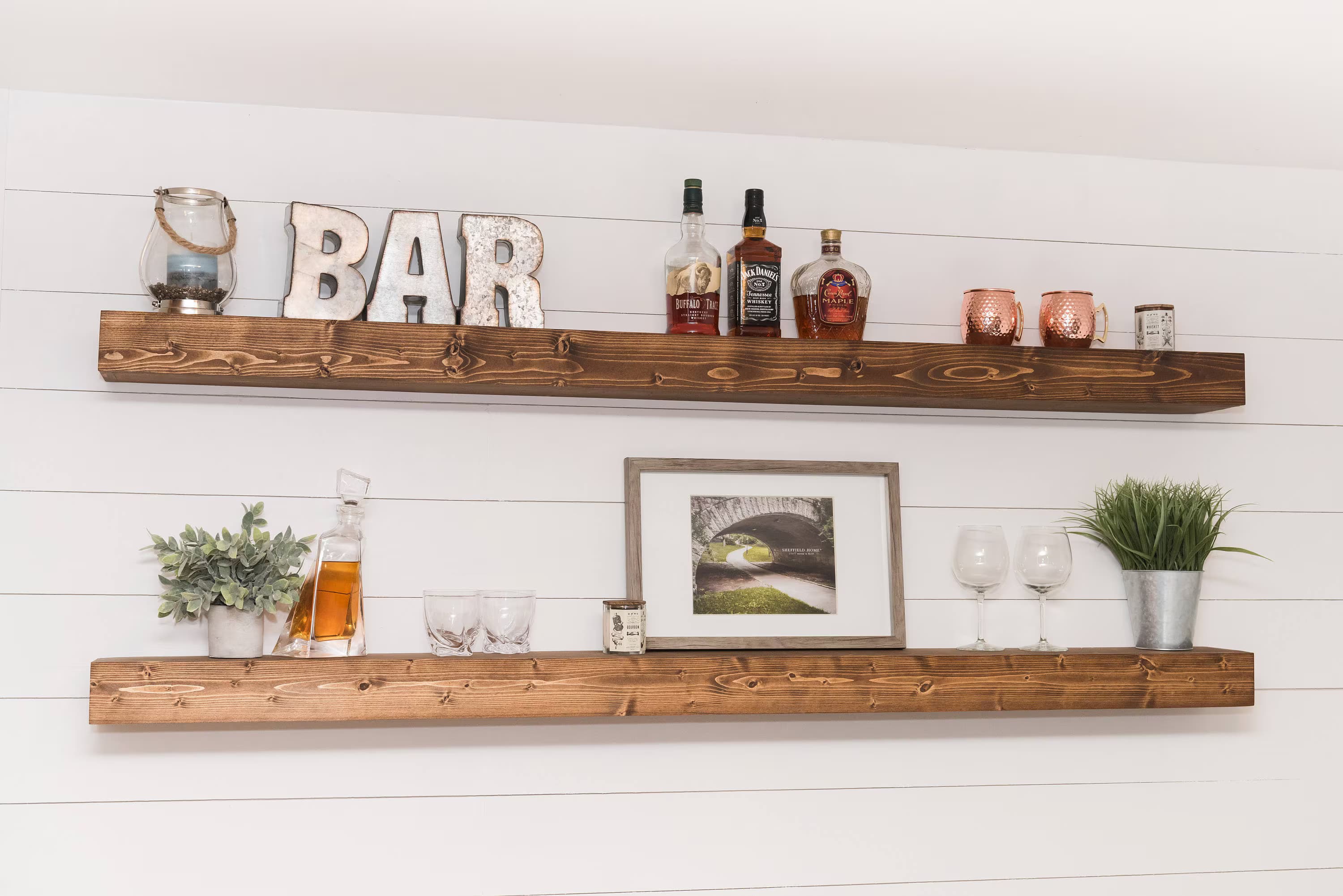 Two large wood floating shelves securely mounted on a shiplap wall holding bar bottles, glassware and decor