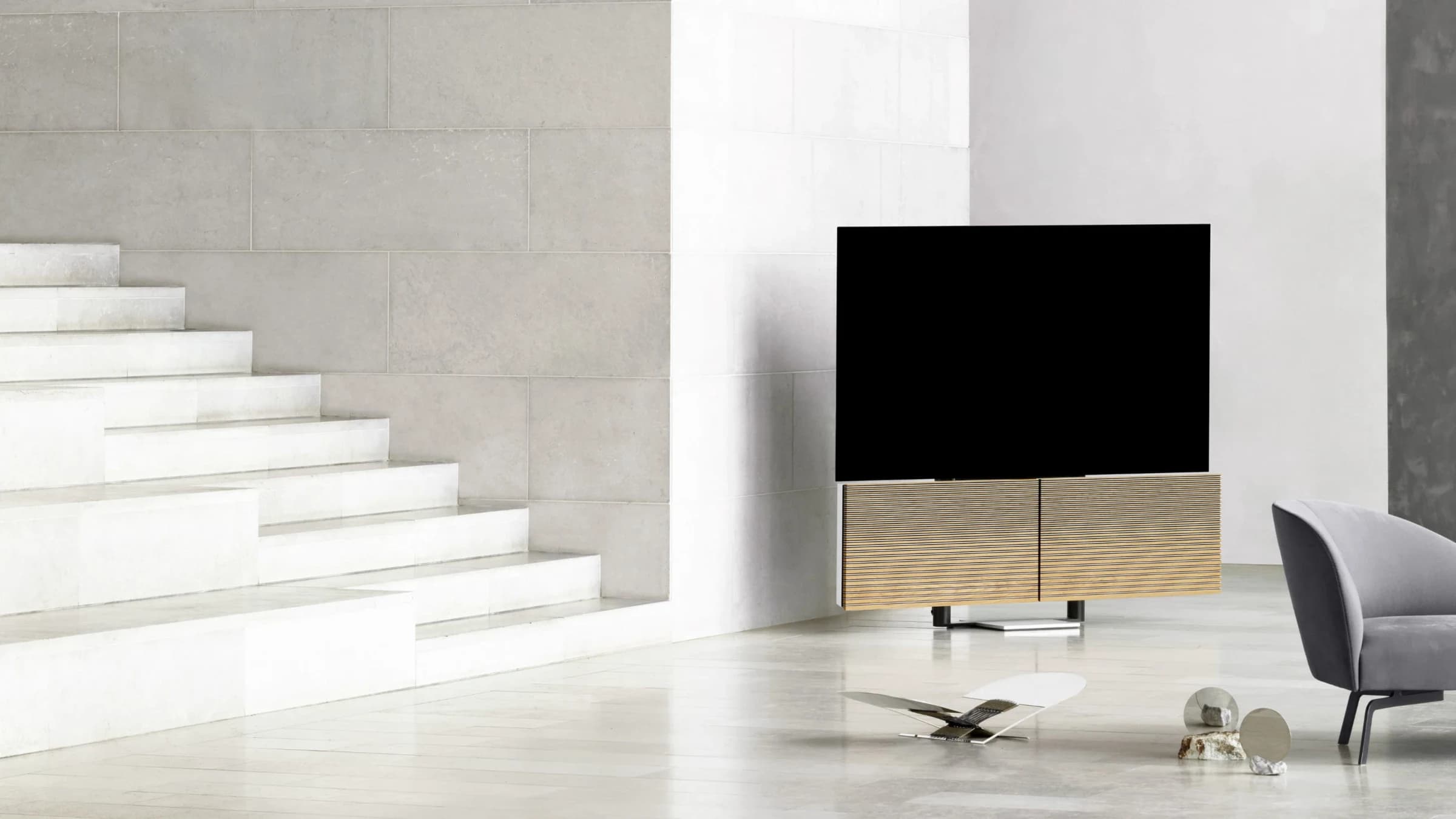 Beovision Harmony television on a floor stand, screen off, placed beside a bright white staircase in a minimal room