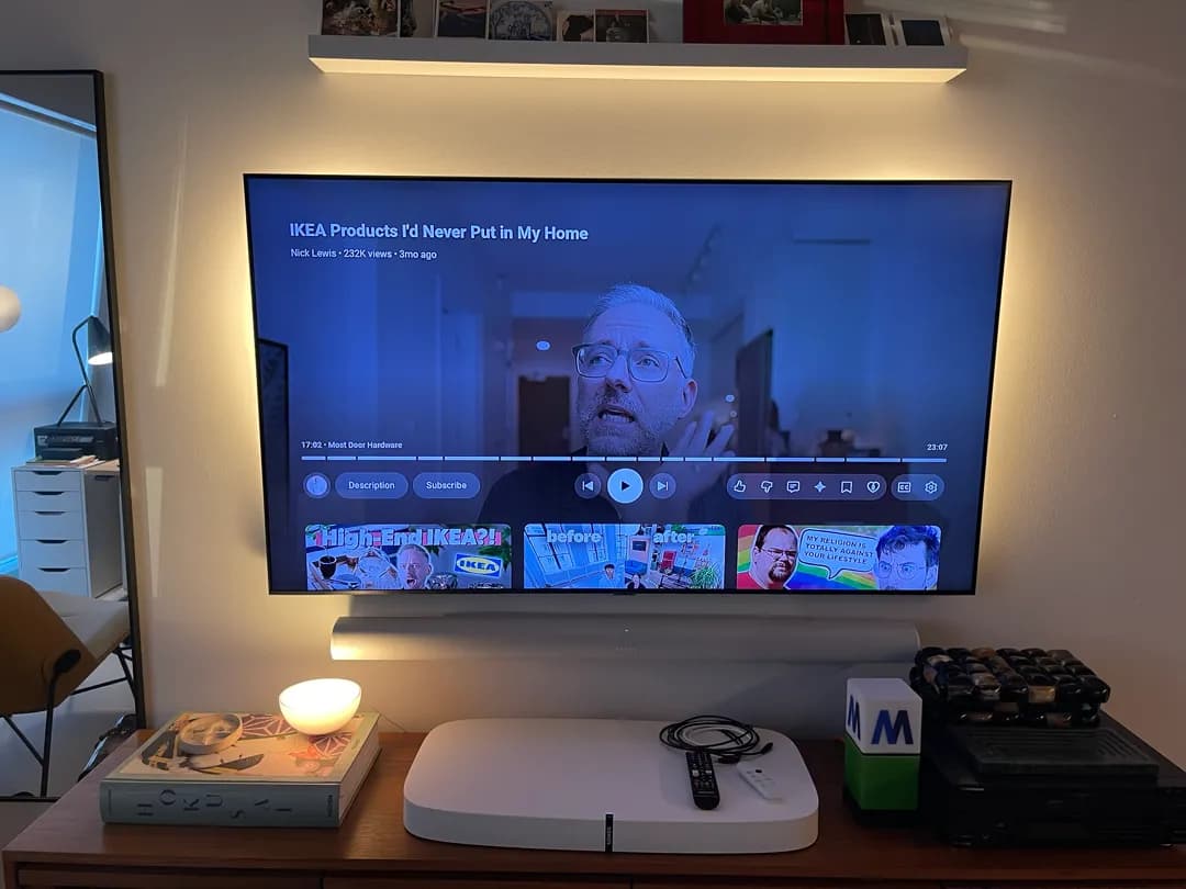 A TV mounted on a wall under a backlit floating shelf plays a video, with a soundbar below and a console holding decor, a sma