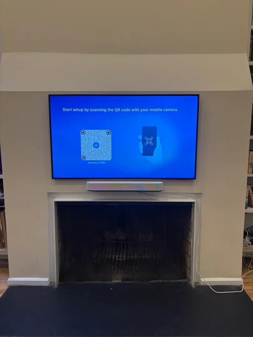 A TV showing a setup screen hangs flat on the chimney wall above a fireplace with a white soundbar beneath it.