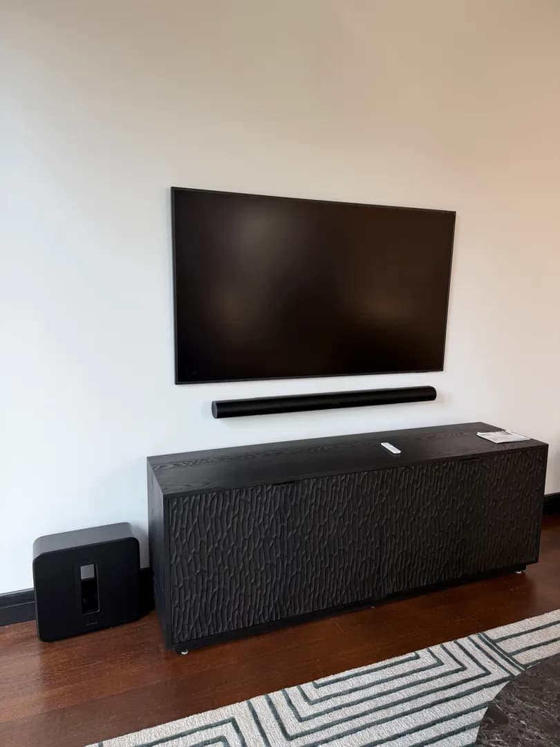 TV and soundbar are mounted on a wall above a black console, with a Sonos subwoofer on the floor beside it.
