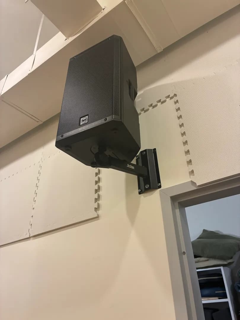 A black powered speaker on a wall bracket beside a doorway, with foam tiles on the wall.