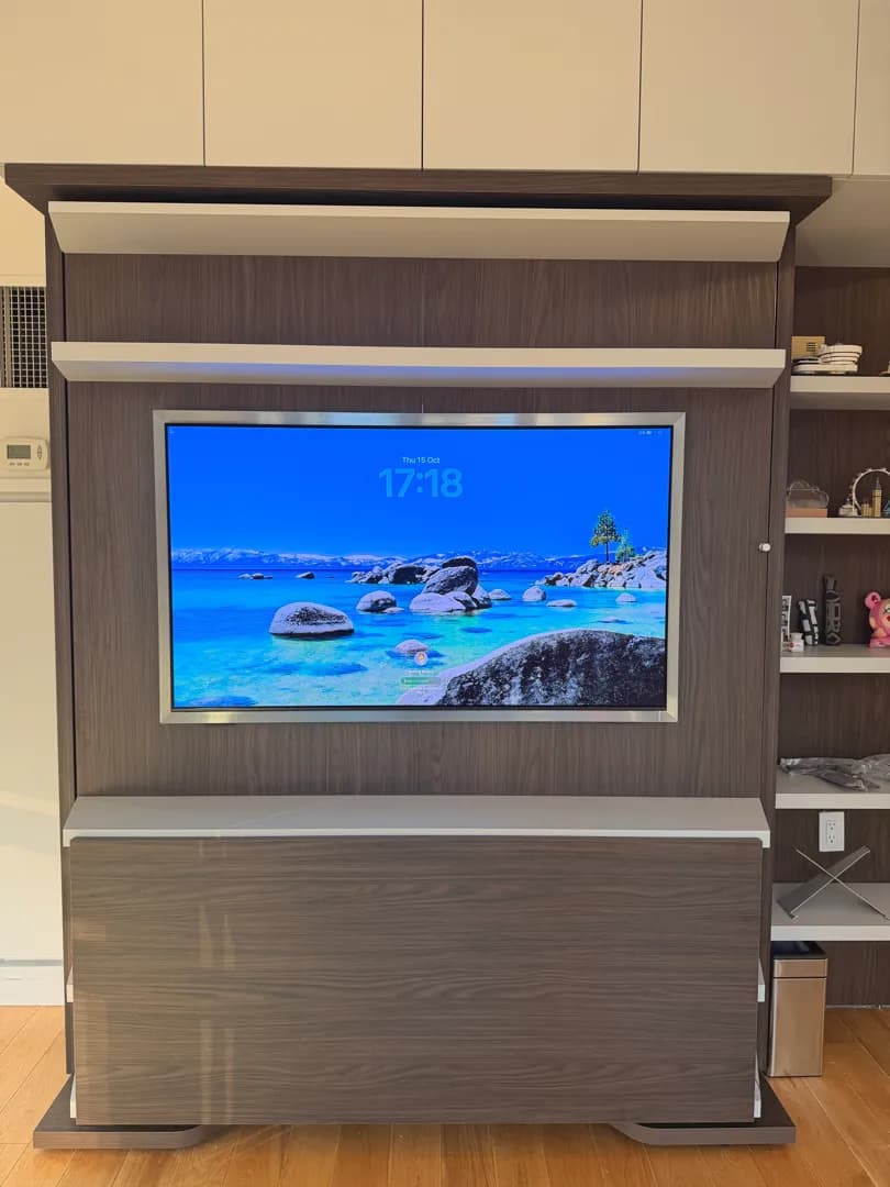 A TV showing a lock screen is mounted inside a wood cabinet unit with a lift mechanism, flanked by open shelving with decor.