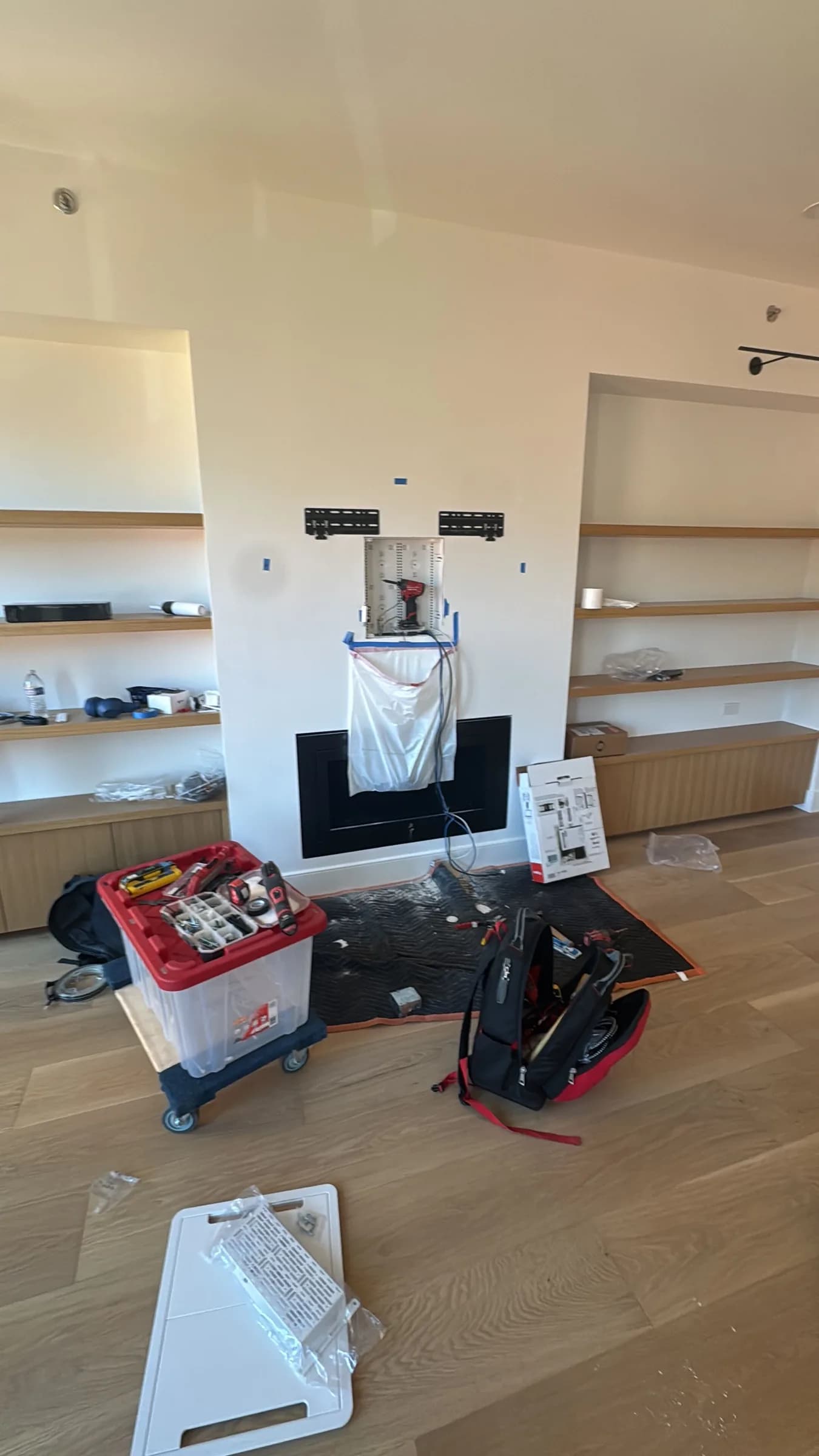 A living room under construction, with a plastic-wrapped fireplace, tool cases, and open wood shelving on both sides.