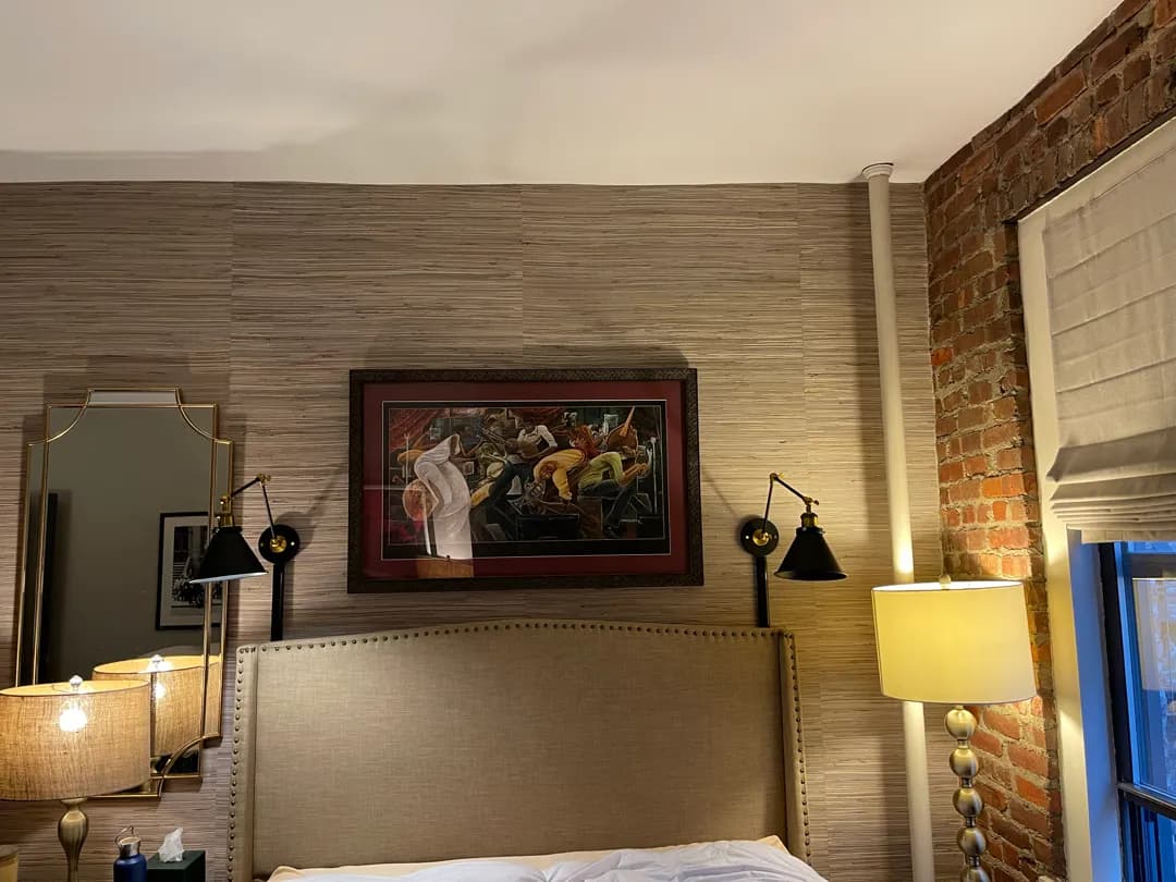A large framed print on a grasscloth wall above a headboard, between two black swing-arm wall lights.