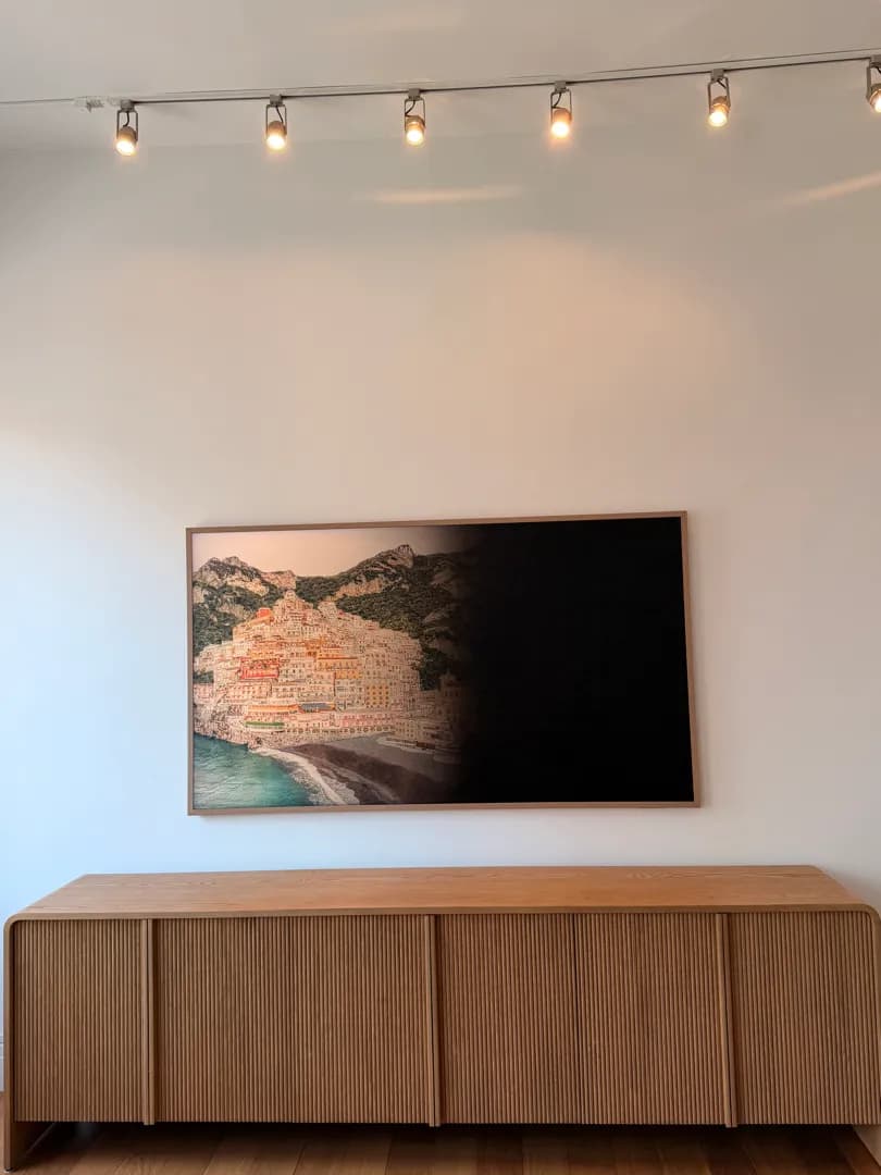A wood-framed TV in art mode displaying a coastal town painting, mounted above a ribbed wood console under track lighting.