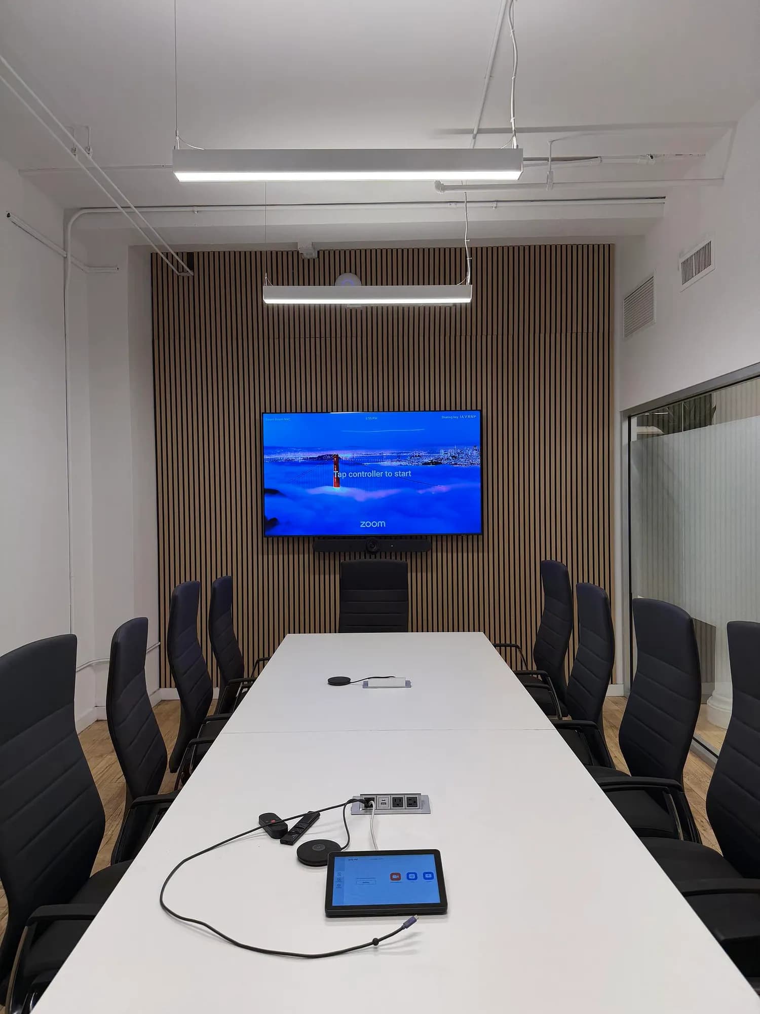 A large boardroom with a wood-slat panel wall behind the TV and camera bar, a long table and black chairs.