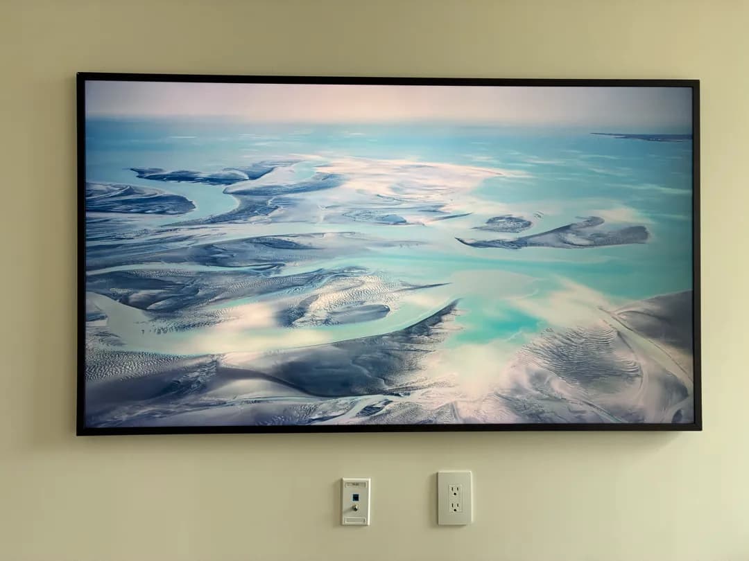 A black-framed Frame TV on a wall showing an aerial sea and sand picture, with a cable plate and an outlet below.
